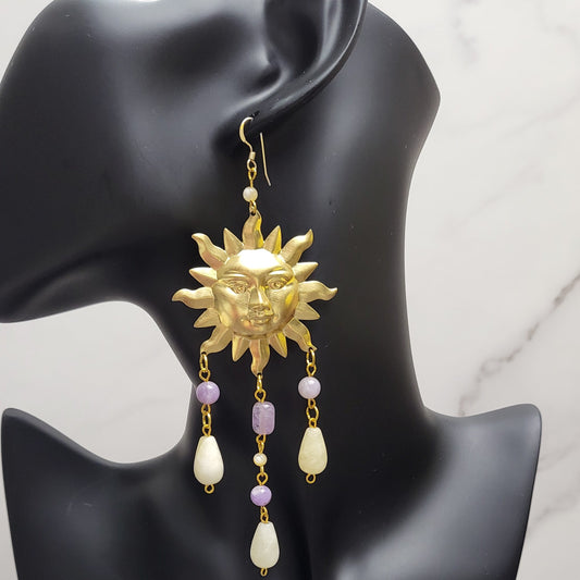 A pair of gold-filled earrings featuring a large sun charm, with vibrant amethyst and earthy tan agate beads, and unbleached mother of pearl accents, displayed on a black mannequin bobbing head.